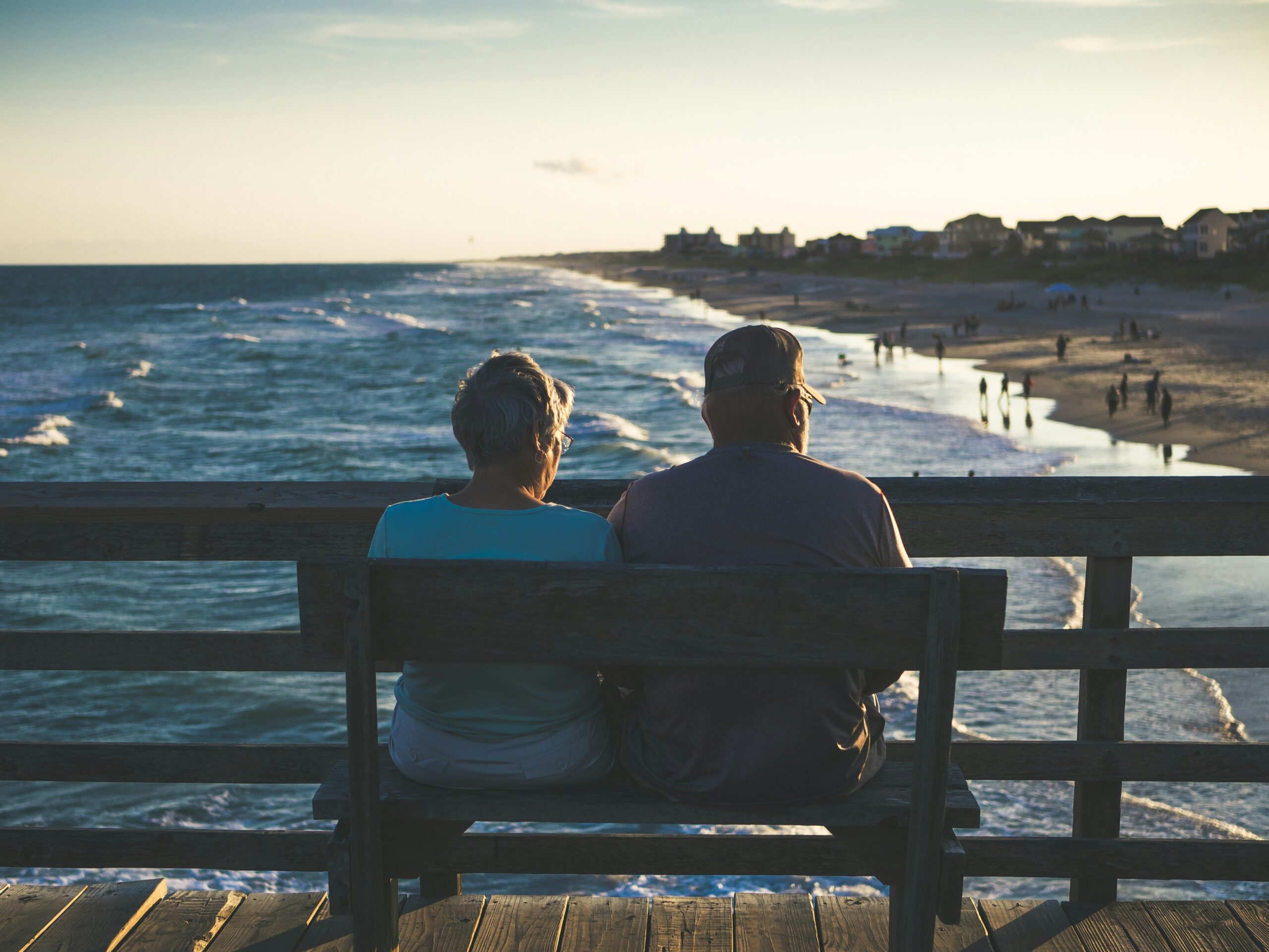An elderly couple sits on a bench looking at a view of the beach.