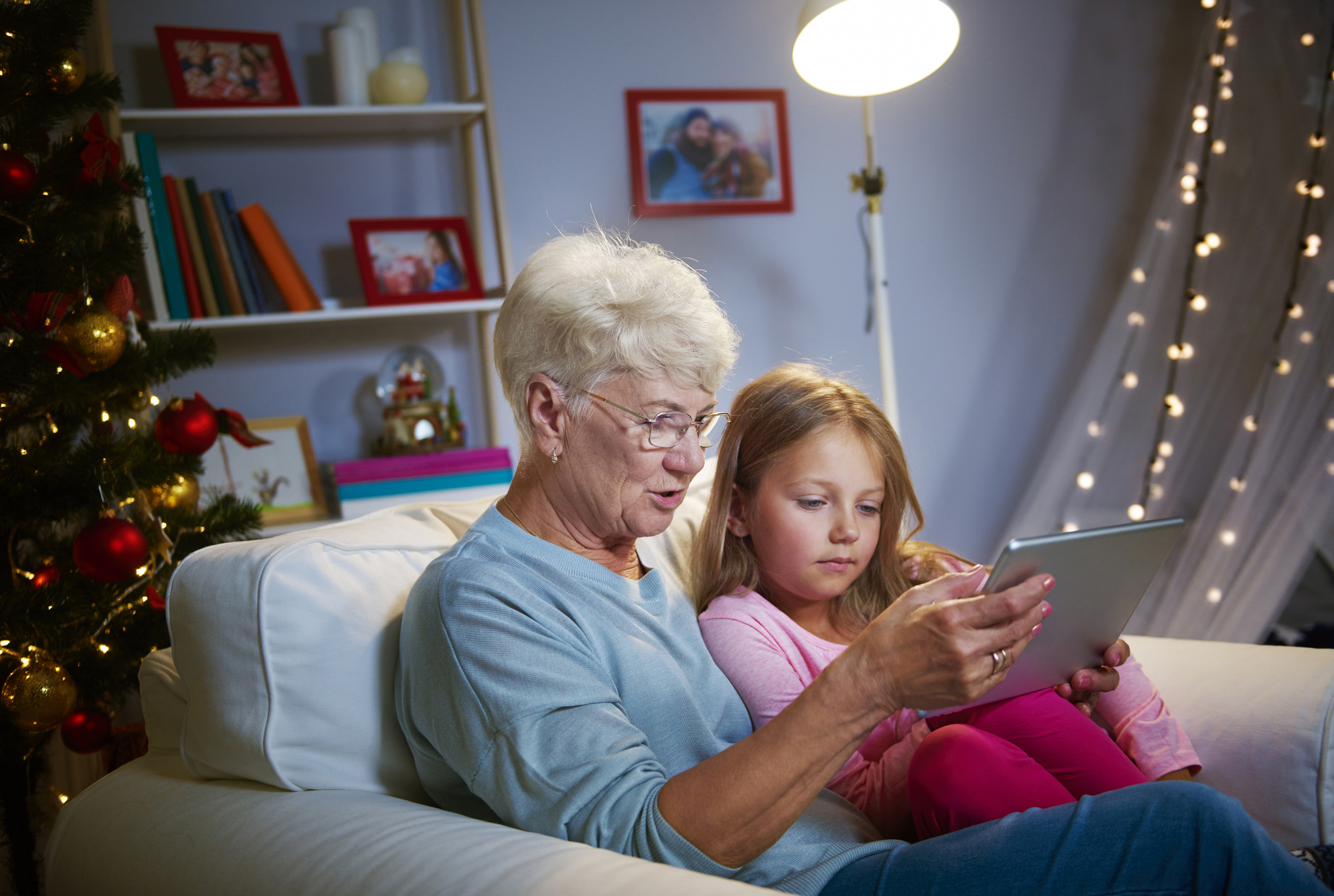 Nanny and little girl sit comfortably on sofa, looking at digital tablet