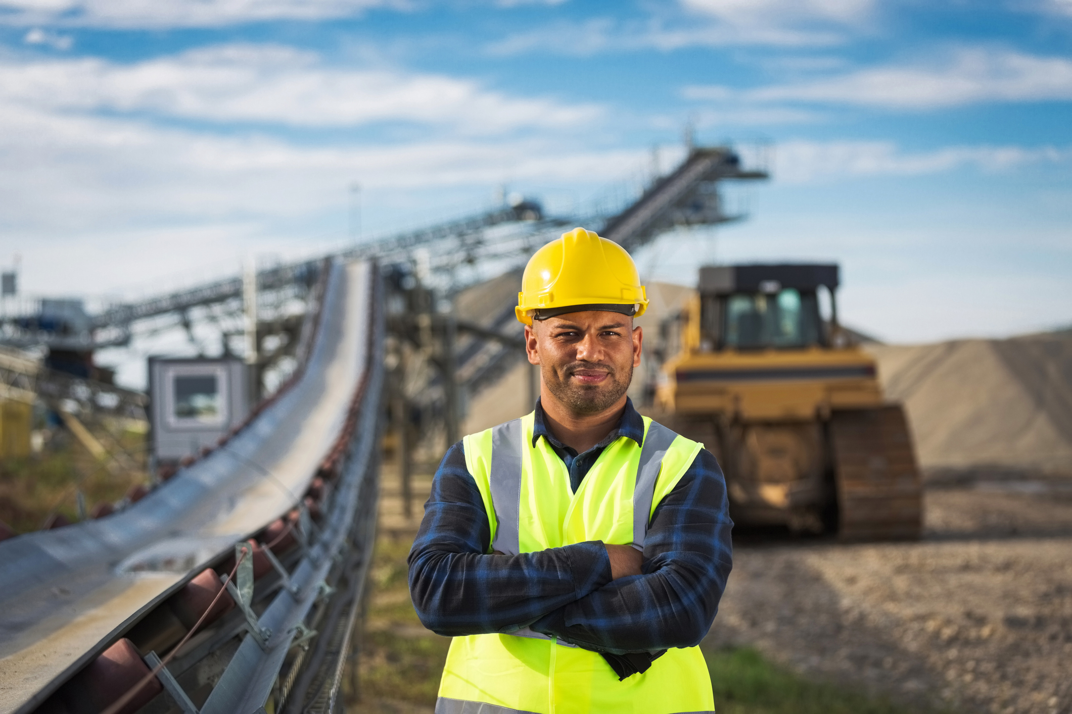 Worker wearing protective clothes and yellow helmet standing next to conveyor belt with arms crossed, smiling at camera.