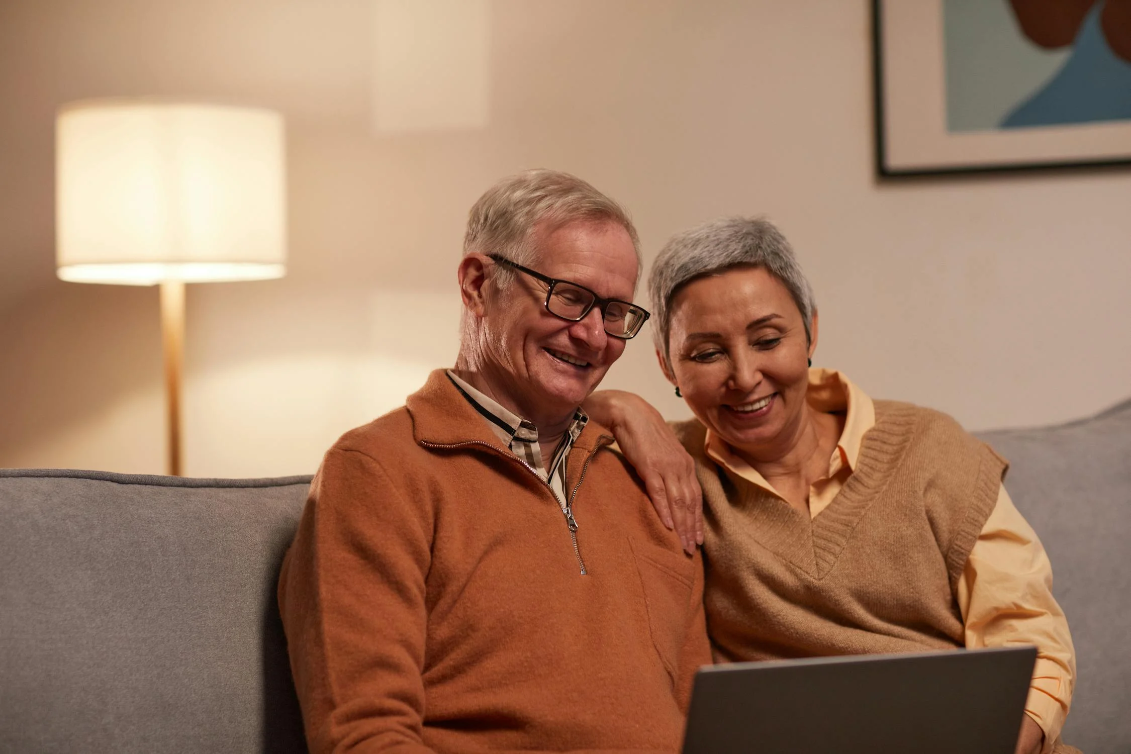 Two older, retired individuals sit on couch contently looking at laptop