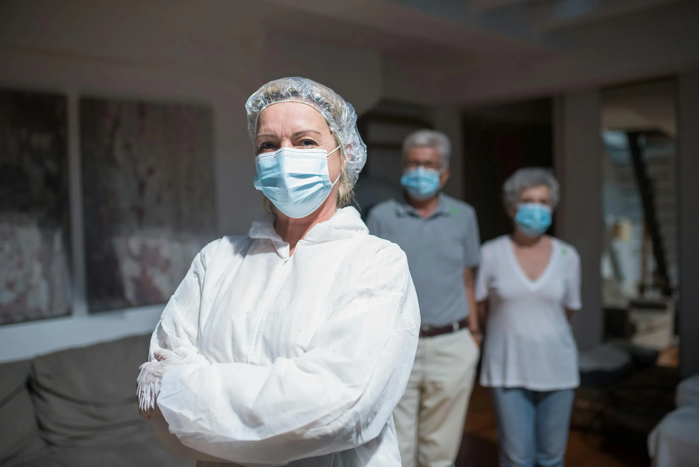 Nurse wearing personal protective equipment stands with her arms crossed, Older couple wearing medical masks stand behind her.