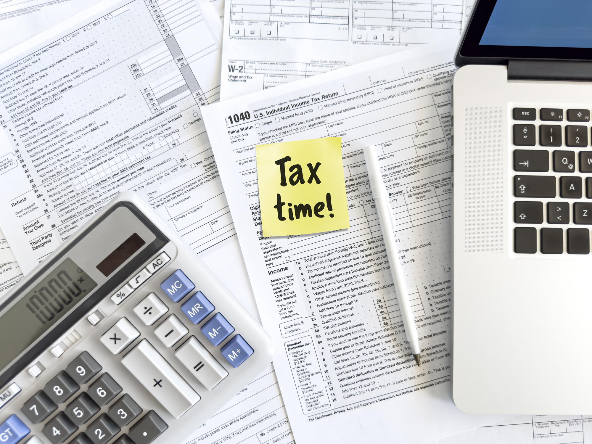 Tax forms, laptop, calculator and adhesive note with “tax time” message with calculator on table top
