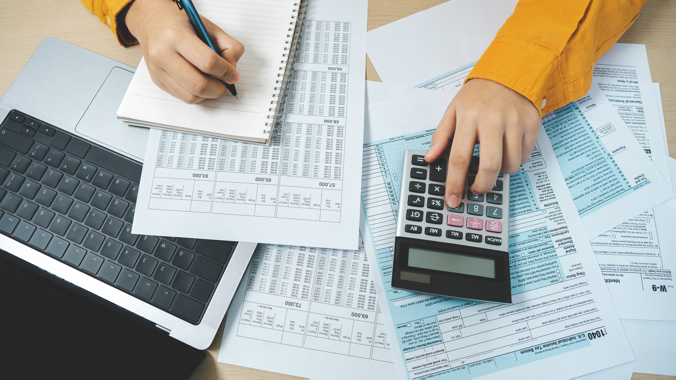 Top view of a person sitting at a desk with tax return preparation and financial management documents, calculating taxes using a calculator and laptop.