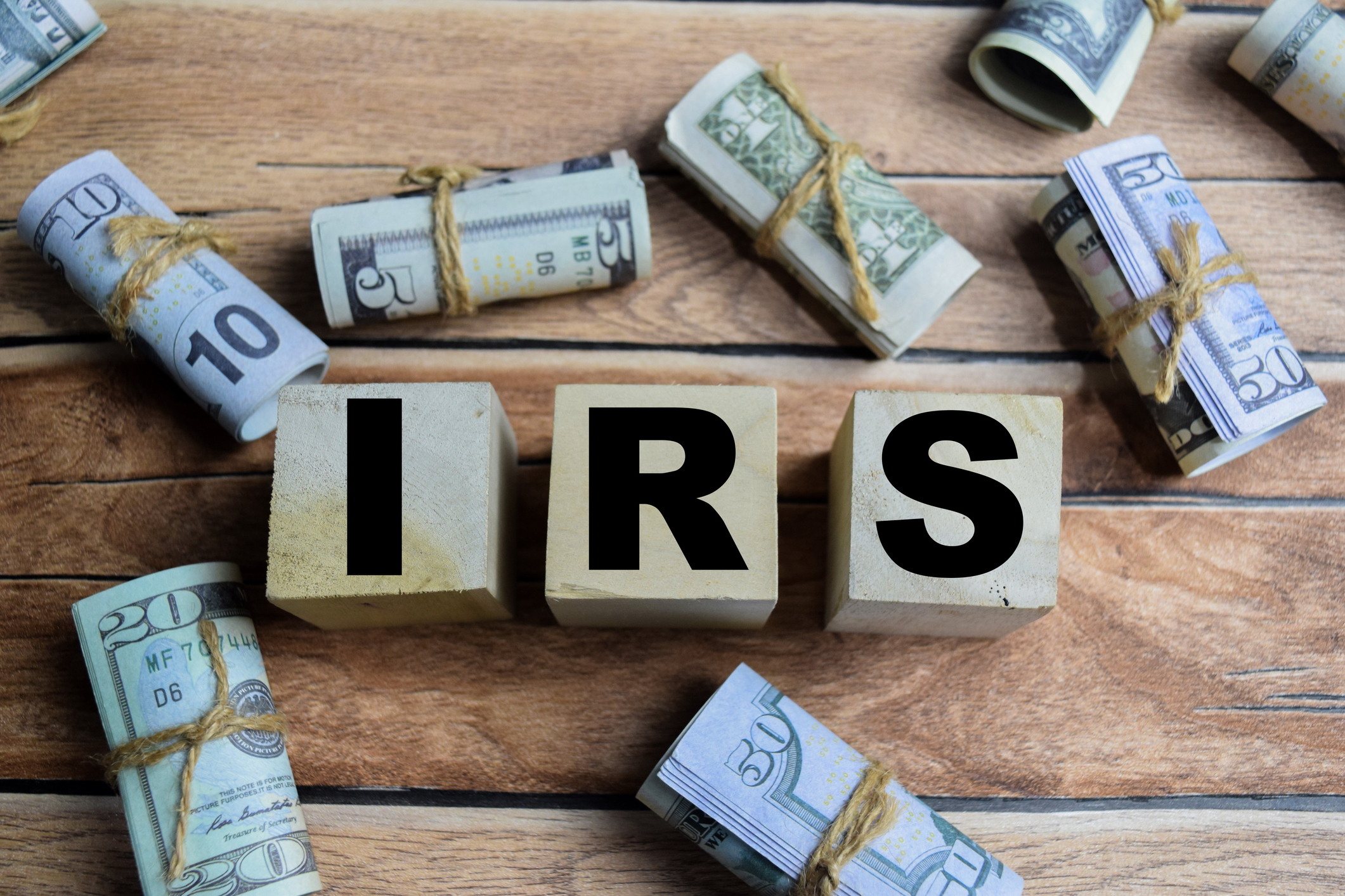 Wooden blocks with the letters "IRS" painted on them rest on a table with US cash currency rolled up in rubber bands.